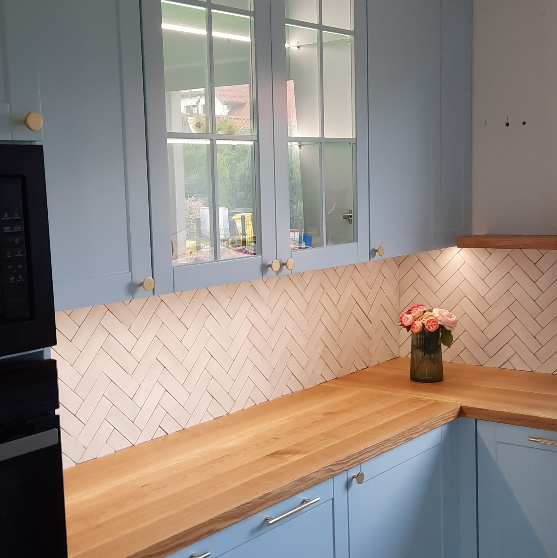 Błękitne szafki kuchenne z drewnianym blatem i dekoracyjną ścianą w jodełkę we Wrocławiu. Na blacie stoi wazon z różowymi kwiatami.