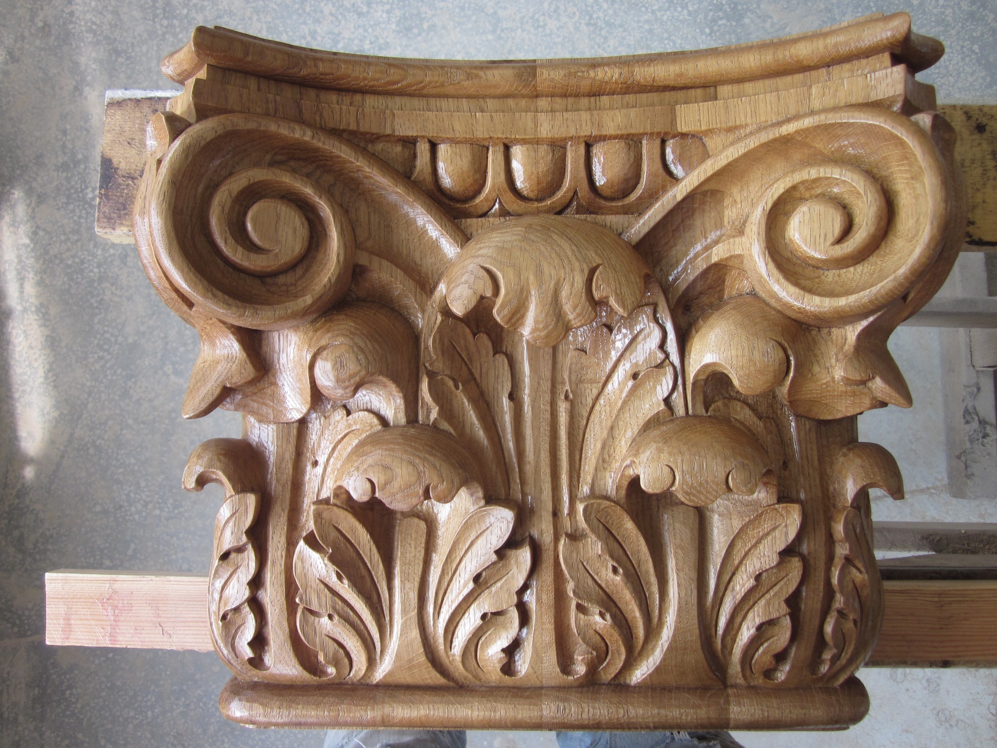 Ręcznie rzeźbiona, dębowa głowica kolumny w stylu korynckim, z wyraźnymi detalami liści akantu i wolut, leżąca na warsztacie, ukazująca kunszt stolarski.