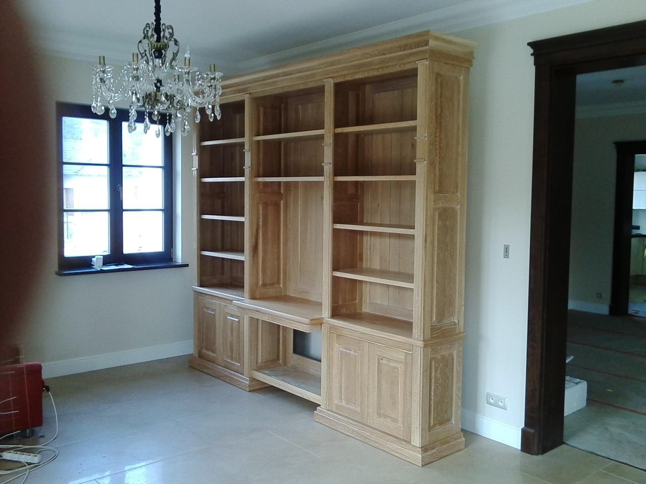 Drewniana biblioteczka z szafkami i półkami, oświetlona przez żyrandol i światło z okna. Widoczny fragment pokoju z podłogą z płytek i ciemną stolarką drzwiową.