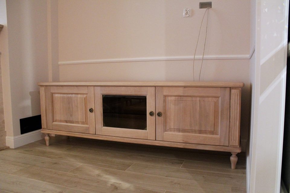 Jasna, drewniana szafka RTV z przeszkloną środkową częścią, ustawiona na podłodze z płytek imitujących drewno. Widoczne detale frezowania i okucia.