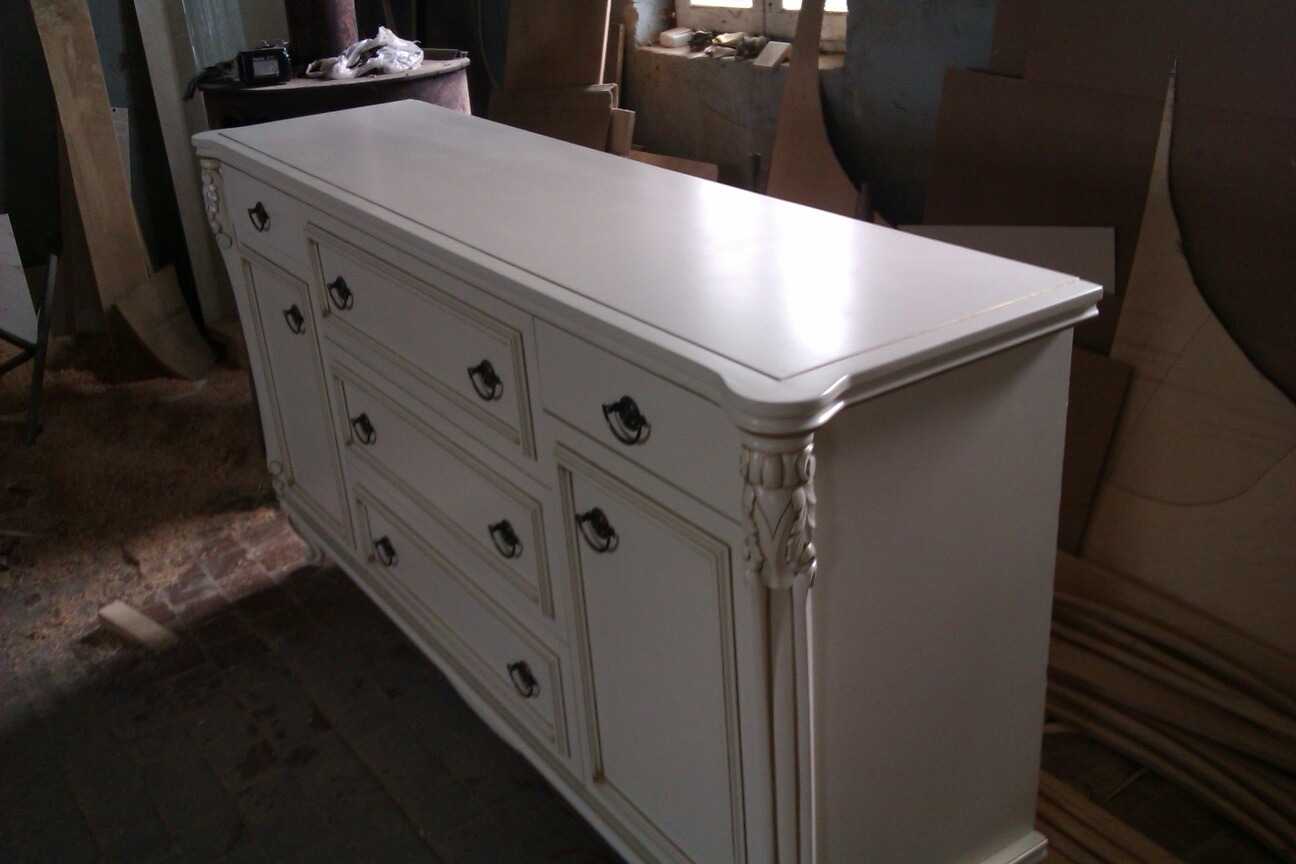 Biała komoda z szufladami, zdobiona ornamentami, w warsztacie stolarskim. Widoczne detale wykończenia i surowe otoczenie.