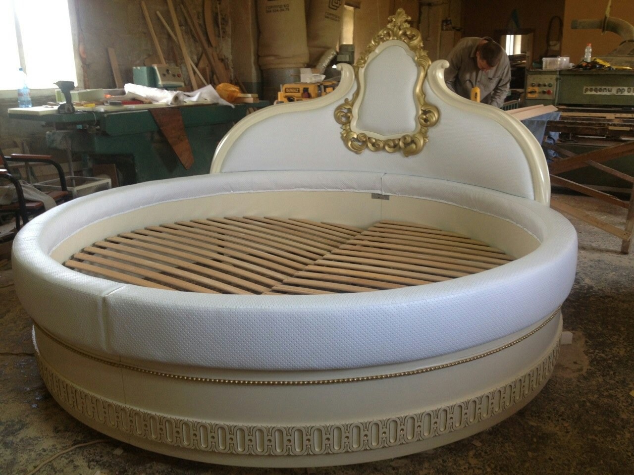 Okrągłe, białe łóżko z drewnianym stelażem i zdobieniami w stylu retro, widoczne w warsztacie stolarskim podczas produkcji.