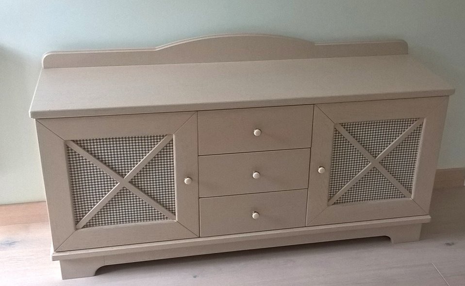 Beżowa komoda z trzema szufladami i szafkami z wzorem w pepitkę, białe gałki, subtelne oświetlenie, drewniana konstrukcja, styl rustykalny.