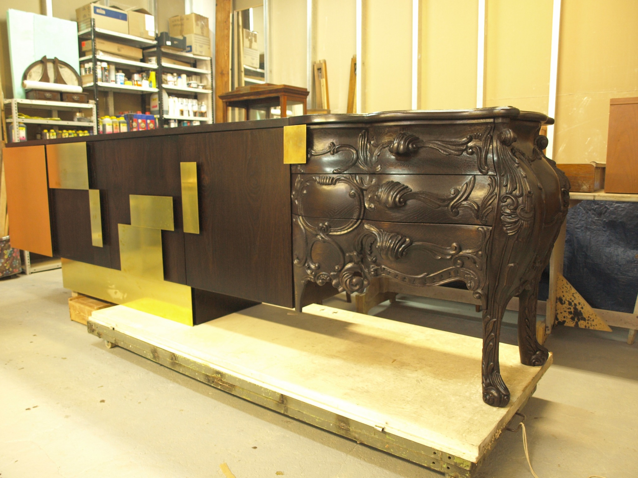 Masywna, ciemna komoda z rzeźbieniami i złotymi elementami geometrycznymi, stojąca na podwyższeniu w warsztacie. Widoczne detale wykończenia i połączenie stylów.