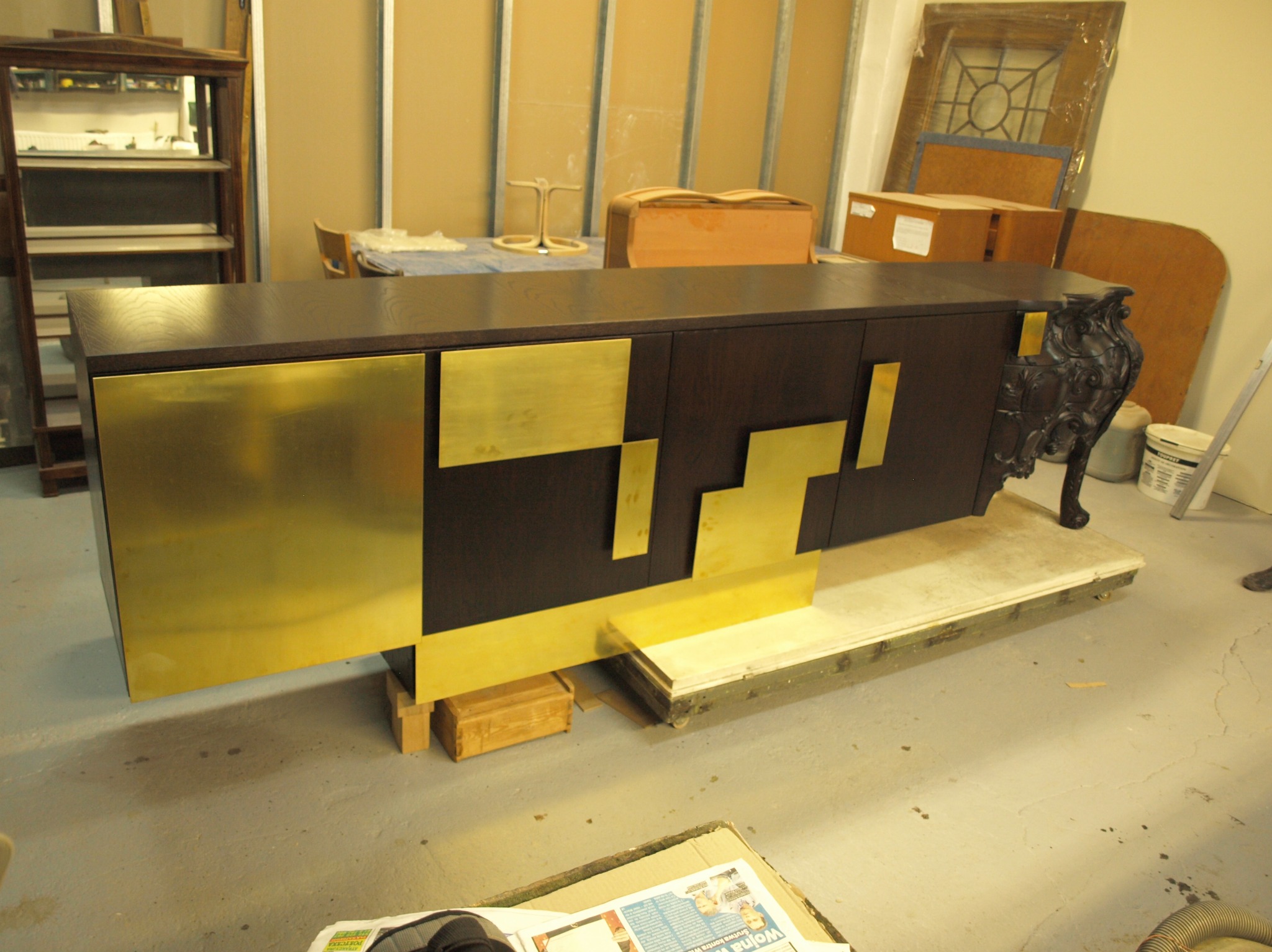 Elegancka komoda z ciemnego drewna z geometrycznymi, złotymi akcentami, wsparta na ozdobnych nogach, w warsztatowym otoczeniu z elementami stolarskimi w tle.