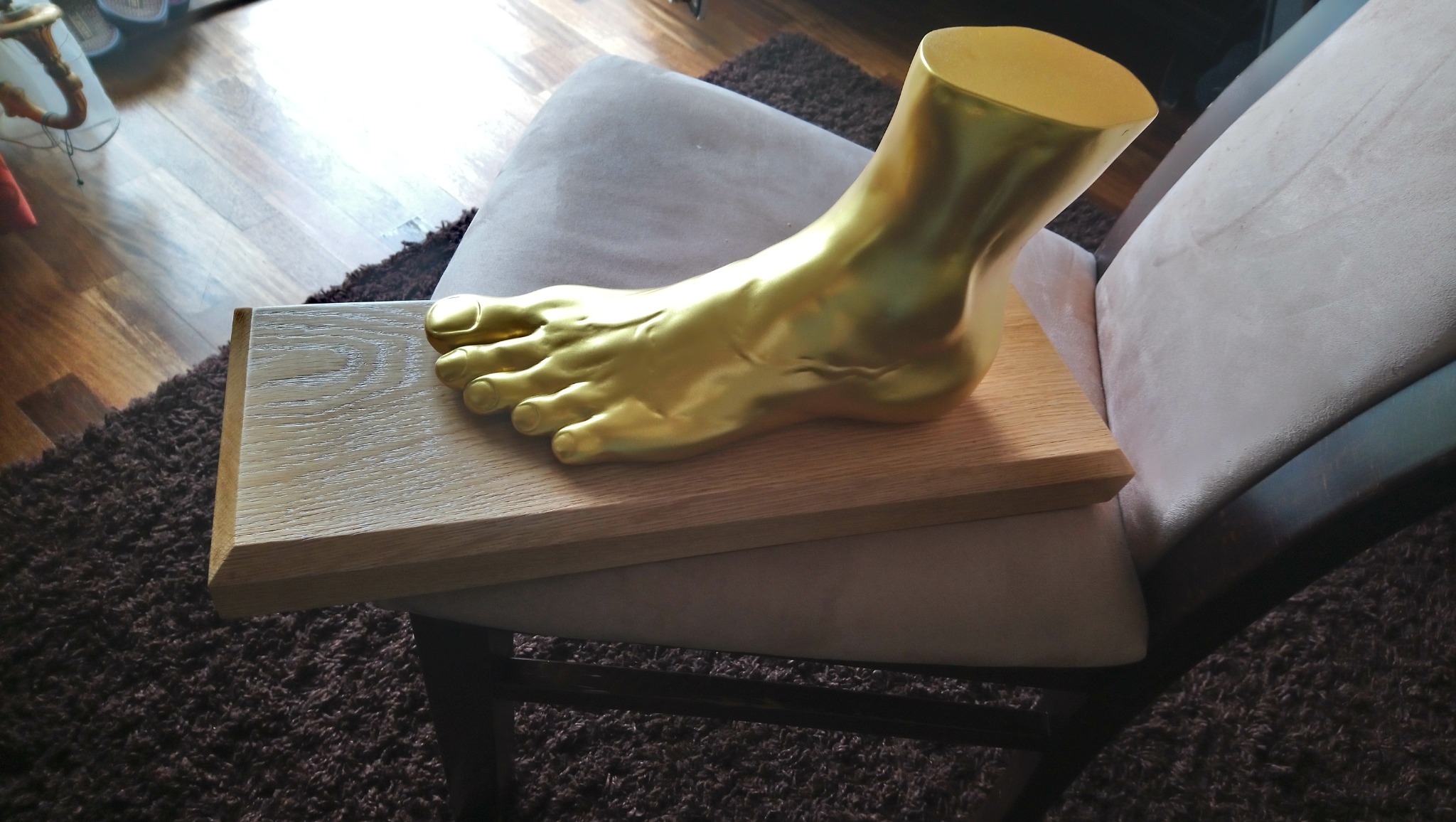 Złota, anatomiczna forma stopy na jasnym, drewnianym blacie, umieszczona na krześle z beżową tapicerką. Ciemny dywan w tle.