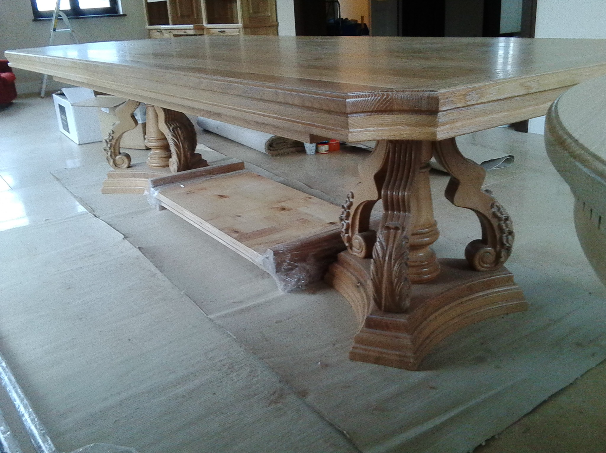 Stylowy, drewniany stół z rzeźbionymi nogami, widoczny w trakcie montażu. Część podłogi zabezpieczona kartonem. W tle jasne meble.