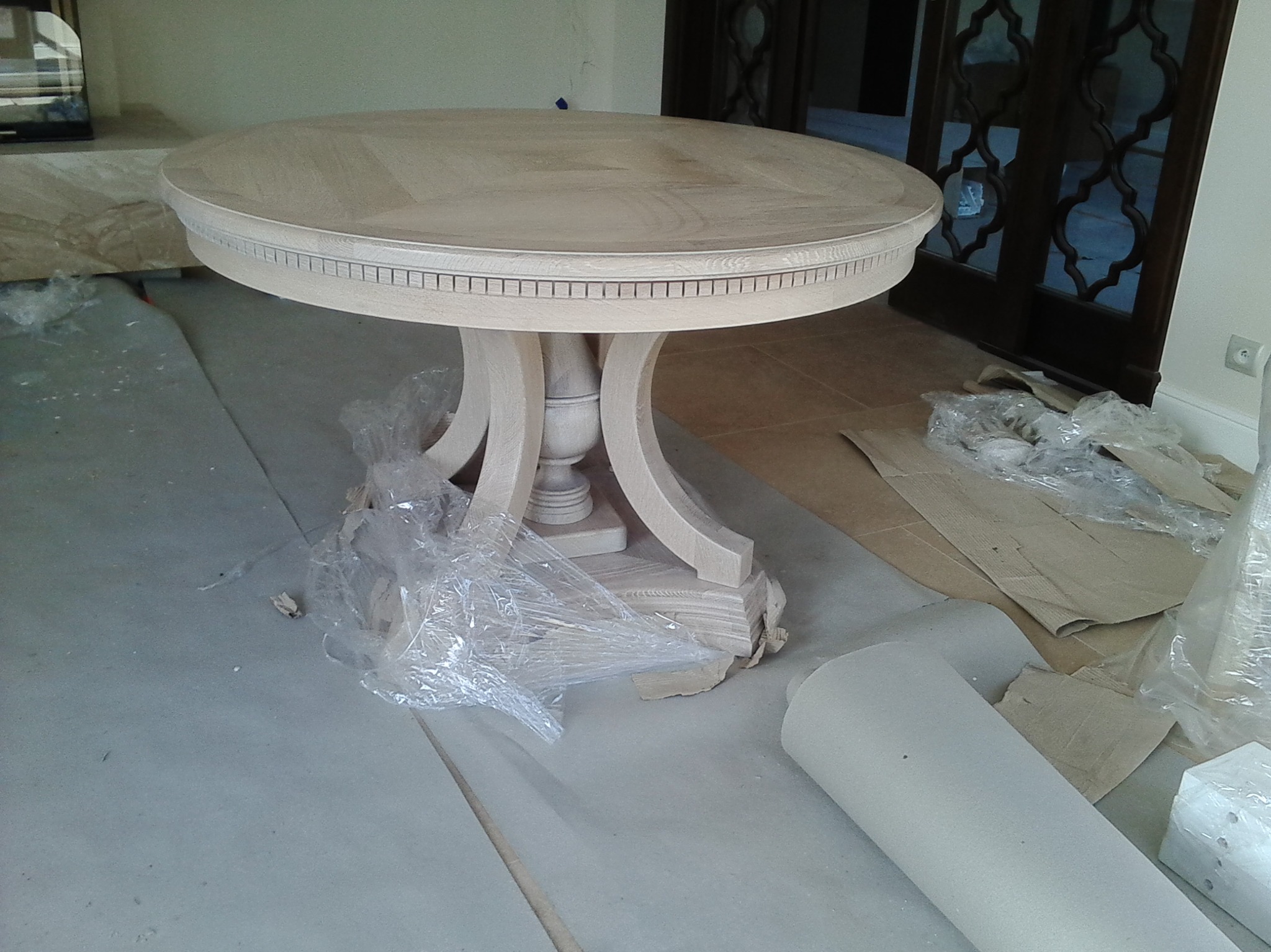 Okrągły, surowy stół drewniany na toczonej nodze, zabezpieczony folią i kartonem, na tle drzwi z dekoracyjnymi szybami. Stolarz.