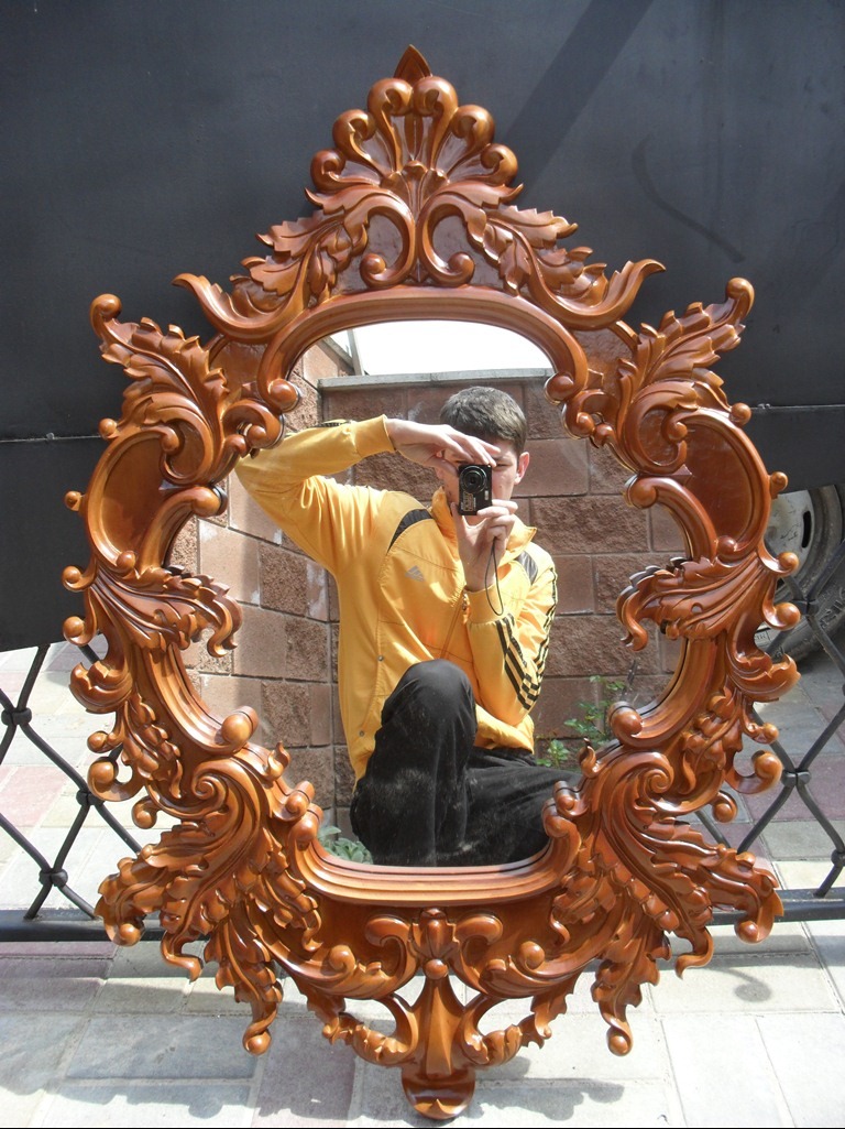 Ozdobne, drewniane lustro w bogato rzeźbionej ramie z motywami roślinnymi, odbijające postać fotografa z aparatem na tle ceglanej ściany i metalowej balustrady.