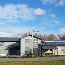 Budynek firmy z dużym niebieskim logo na szarej fasadzie, panele słoneczne na dachu i banery reklamowe przy ogrodzeniu.