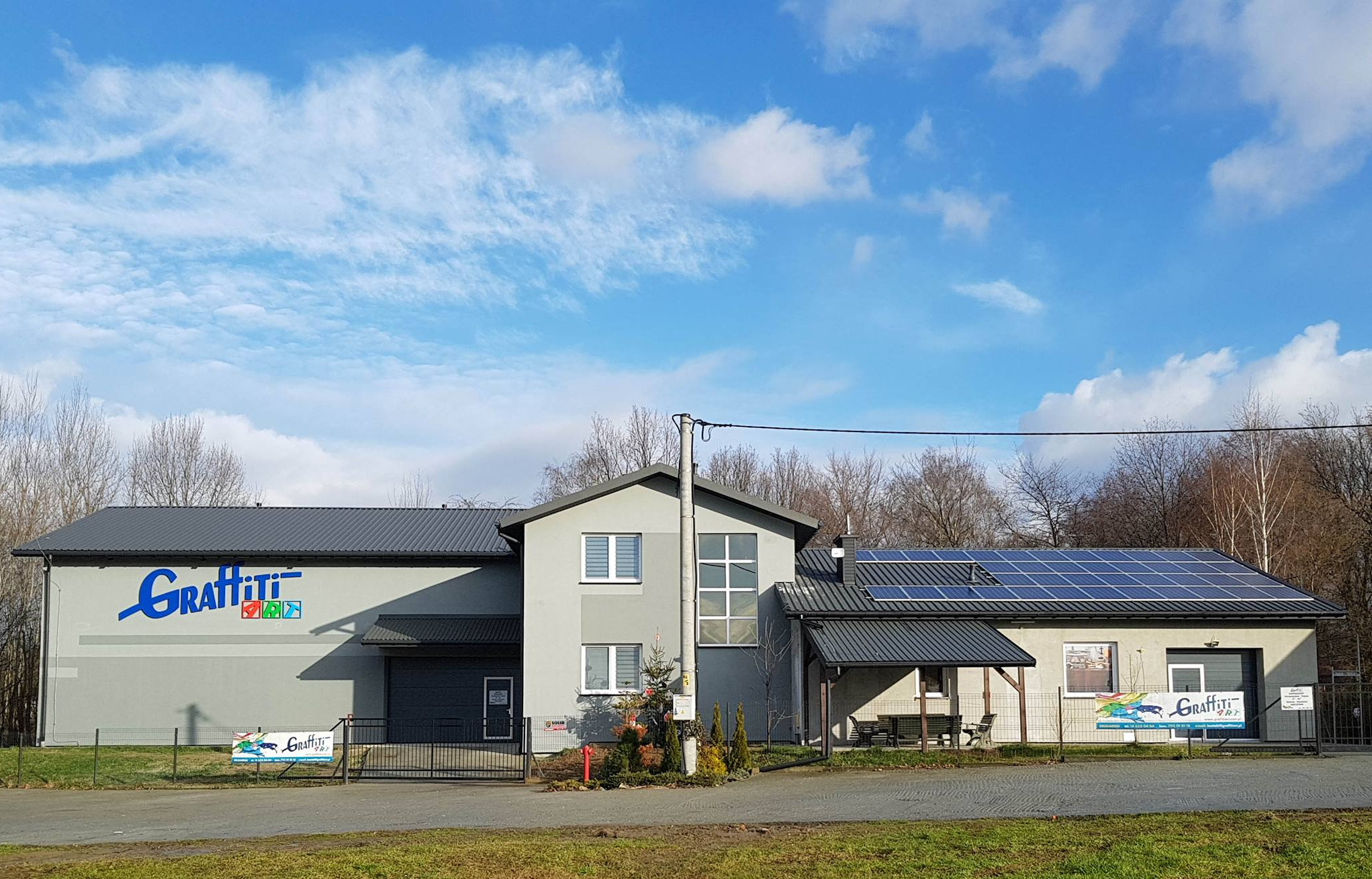 Budynek firmy z dużym niebieskim logo na szarej fasadzie, panele słoneczne na dachu i banery reklamowe przy ogrodzeniu.