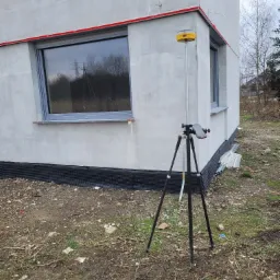 Żółty odbiornik GNSS na statywie, z zamontowanym smartfonem, ustawiony przed surową elewacją szarego budynku z czarnymi oknami i czerwoną listwą, na grząskim gruncie.