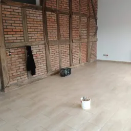 Wykończone wnętrze z ułożoną jasną podłogą z płytek imitujących drewno, ściana z cegły i drewnianych belek, wiadro z farbą na podłodze, czarna kurtka wisząca na ścianie.