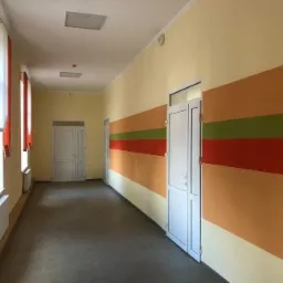 Pomalowany korytarz w budynku użyteczności publicznej z poziomymi pasami w kolorach pomarańczowym, czerwonym i zielonym, białe drzwi i grzejniki, pomarańczowe pionowe żaluzje w oknach.