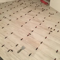 Układanie jasnych płytek imitujących drewno z użyciem systemu poziomowania, widoczna szpachelka do rozprowadzania kleju i czerwona ścierka w rogu pomieszczenia.