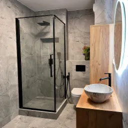 Nowoczesna łazienka z kabiną prysznicową typu walk-in z czarnymi profilami, umywalką nablatową w odcieniach szarości i szafką z drewnopodobnym fornirem oraz okrągłym lustrem z podświetleniem LED.