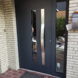 Nowoczesne, antracytowe drzwi wejściowe z pionowym, srebrnym uchwytem i bocznym oknem, zamontowane w ścianie z jasnej cegły, widoczne z zewnątrz na tle brukowanej powierzchni.