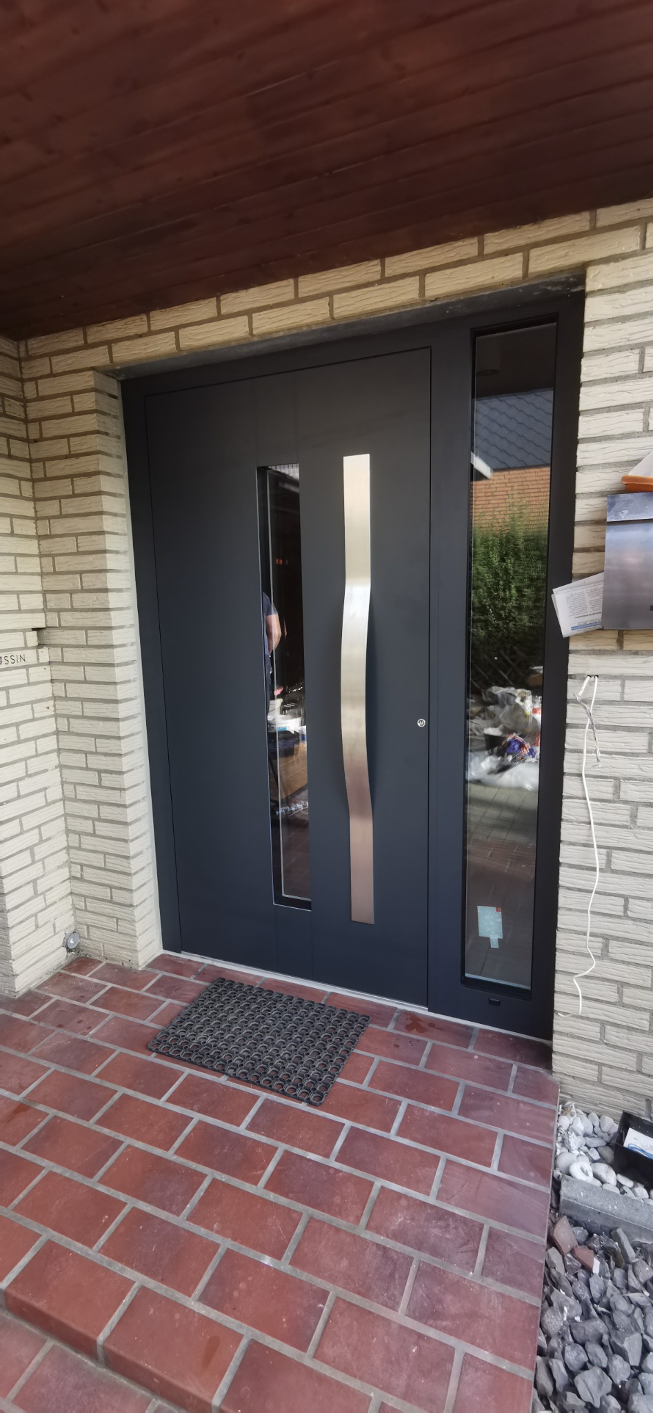 Nowoczesne, antracytowe drzwi wejściowe z pionowym, srebrnym uchwytem i bocznym oknem, zamontowane w ścianie z jasnej cegły, widoczne z zewnątrz na tle brukowanej powierzchni.