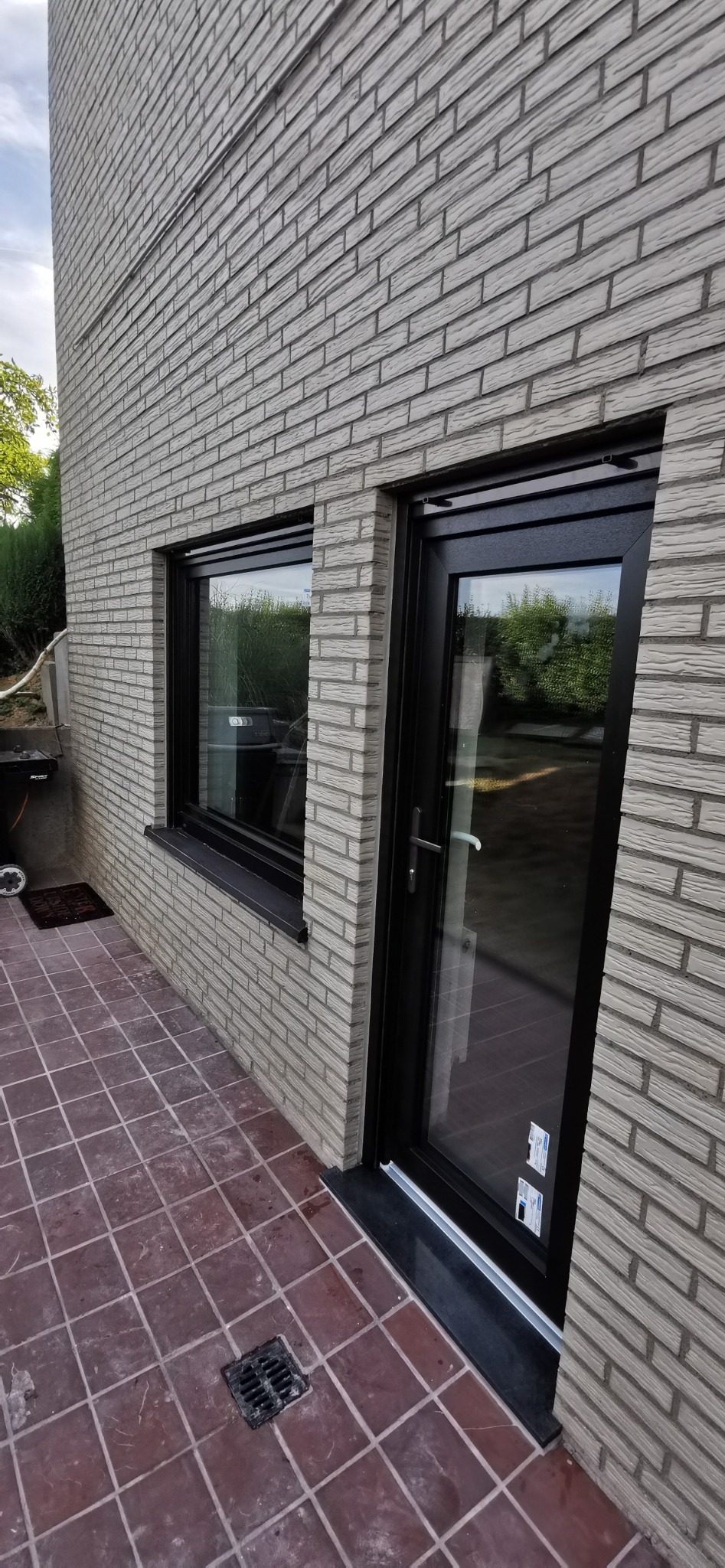 Nowoczesny budynek z szarej cegły z czarną ramą okienną i szklanymi drzwiami, widok z zewnątrz na brązową terakotę.