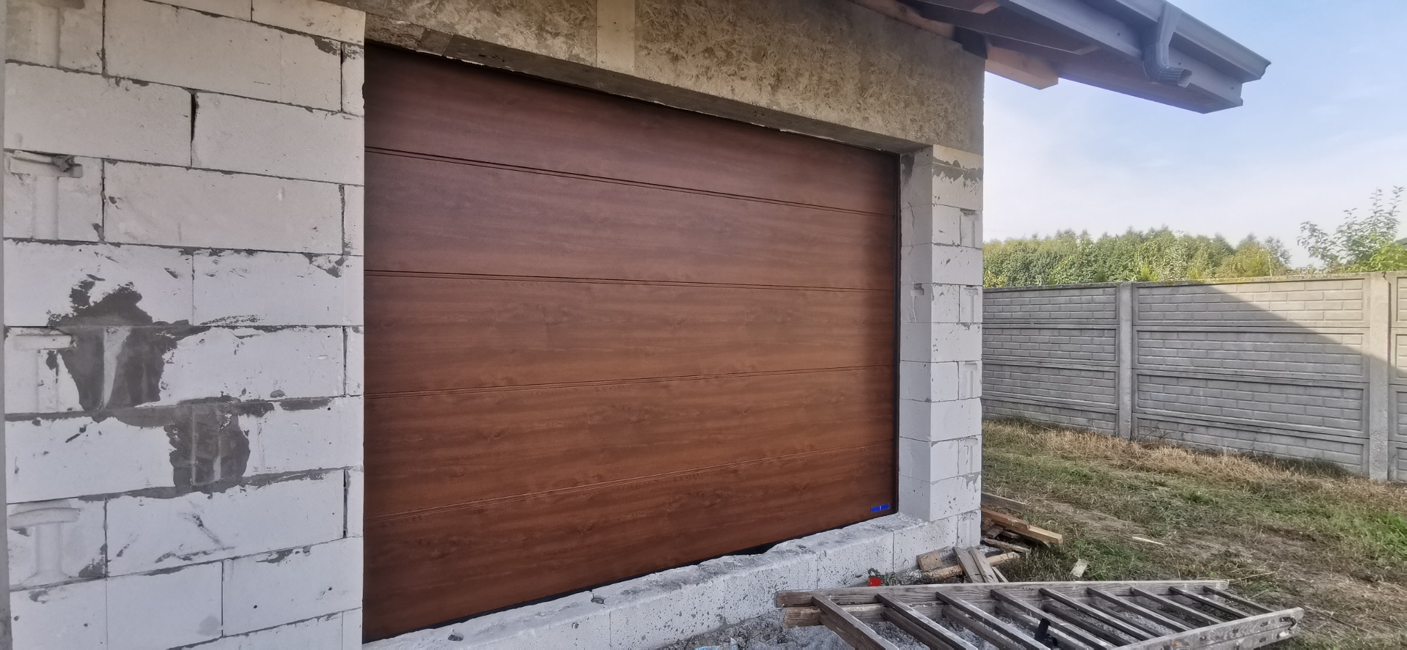Brązowa segmentowa brama garażowa zamontowana w surowym otworze budynku z bloczków betonowych, widok z zewnątrz, drabina oparta o ścianę.