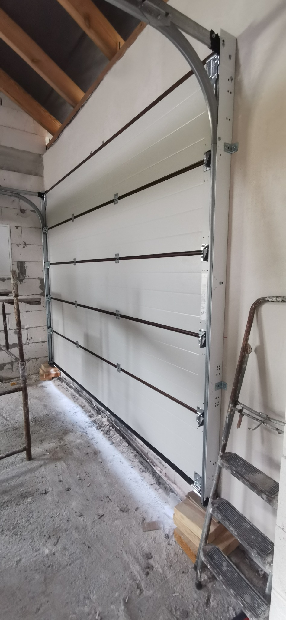 Biała segmentowa brama garażowa z szarymi prowadnicami, widoczna podczas montażu w surowym stanie budynku, z drabiną opartą o ścianę po prawej stronie.
