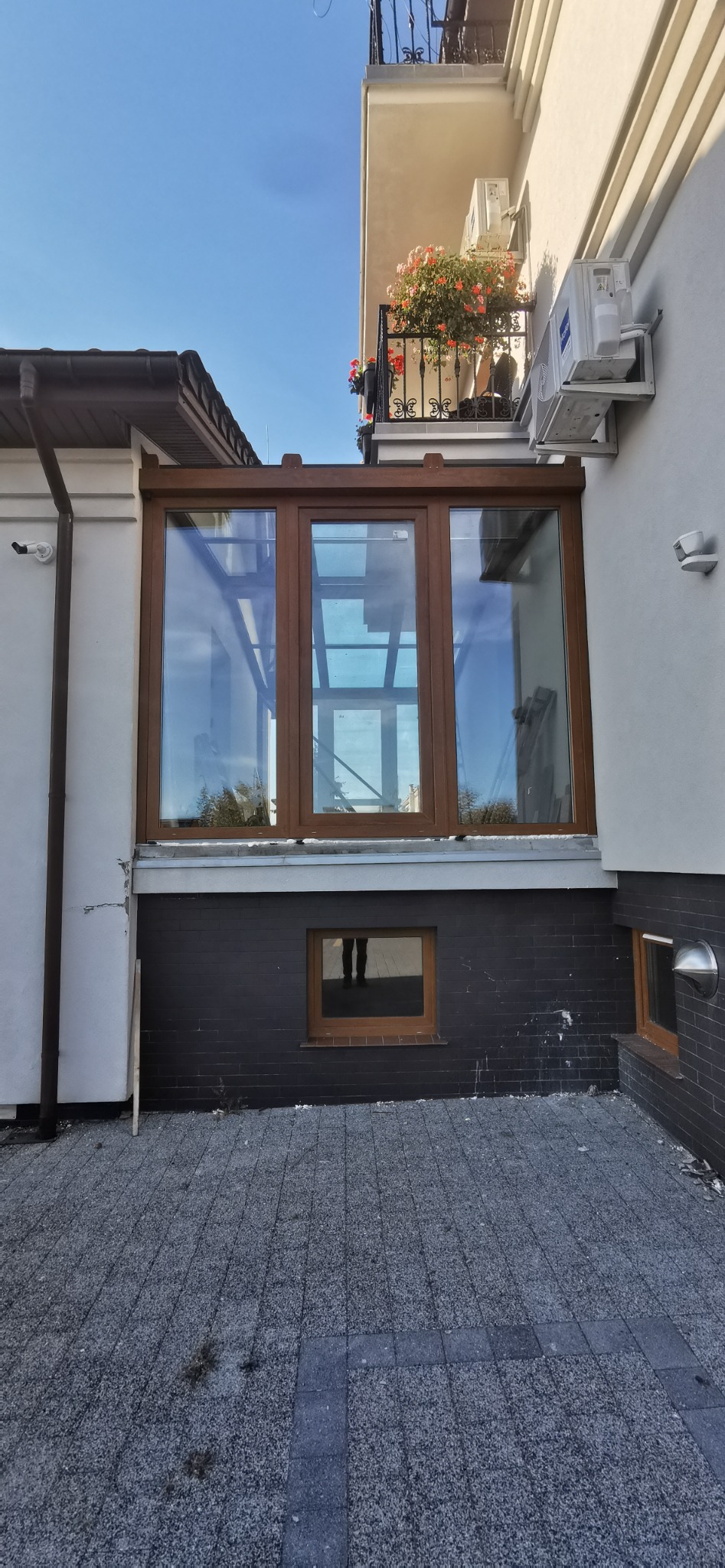 Zewnętrzny widok przeszklonej werandy z oknami PCV w brązowych ramach, z widocznym odbiciem sylwetki w małym okienku piwnicznym i balkonem z kwiatami na górze.