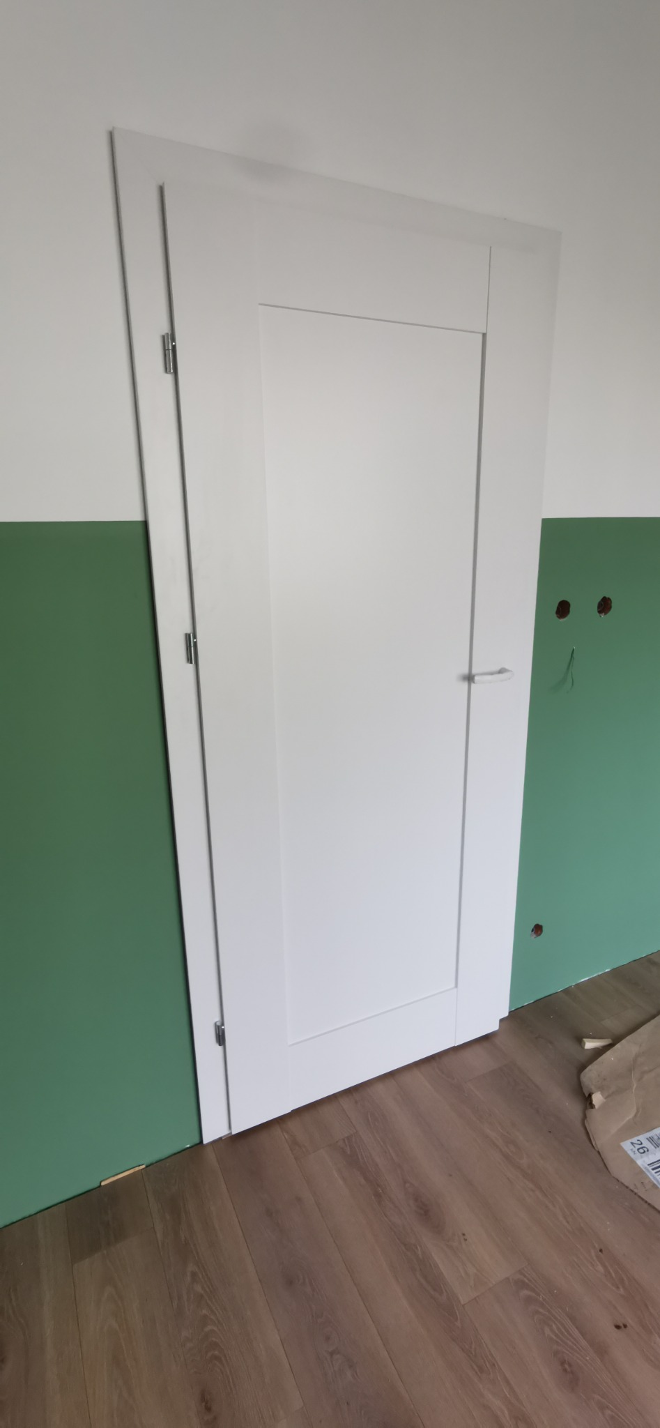 Białe drzwi wewnętrzne z ościeżnicą, zamontowane w pomieszczeniu z zieloną lamperią i drewnopodobną podłogą. Widoczne zawiasy i klamka. Ślady po instalacji elektrycznej na ścianie.