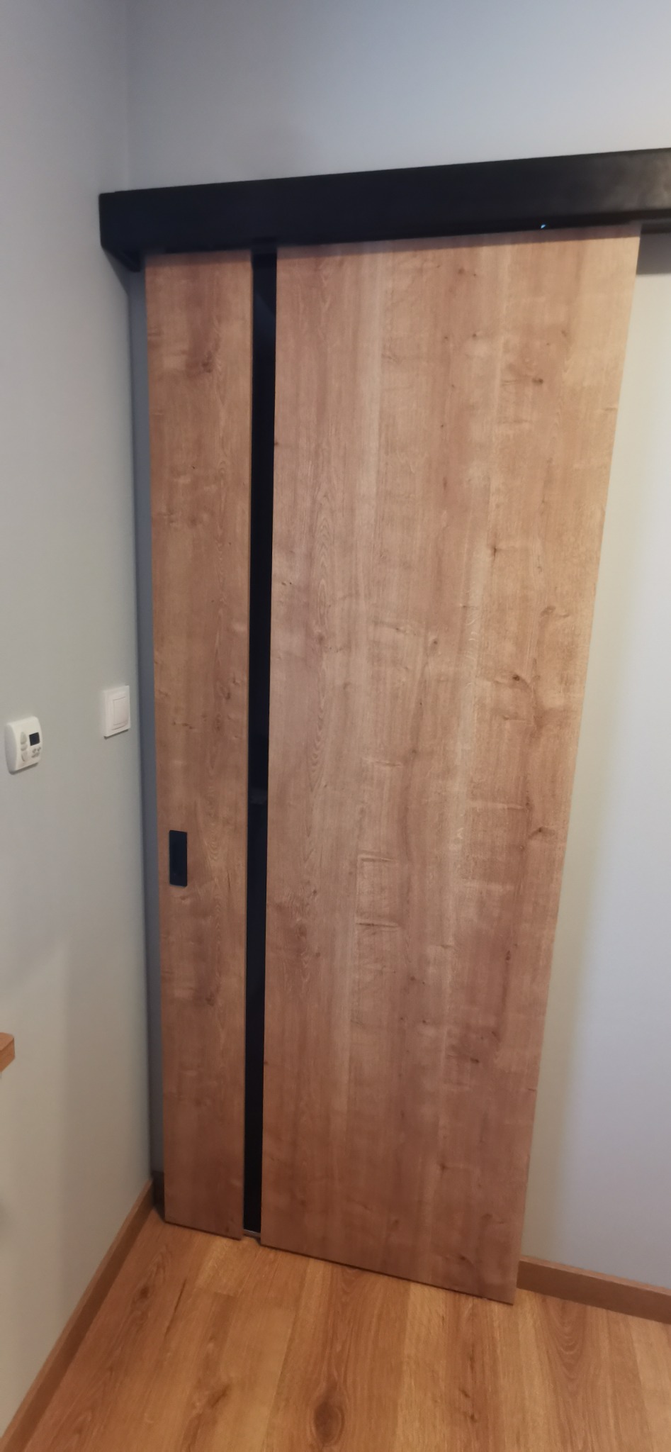 Drewniane drzwi przesuwne z czarnymi akcentami i prowadnicą, zamontowane w jasnym wnętrzu na podłodze imitującej drewno.