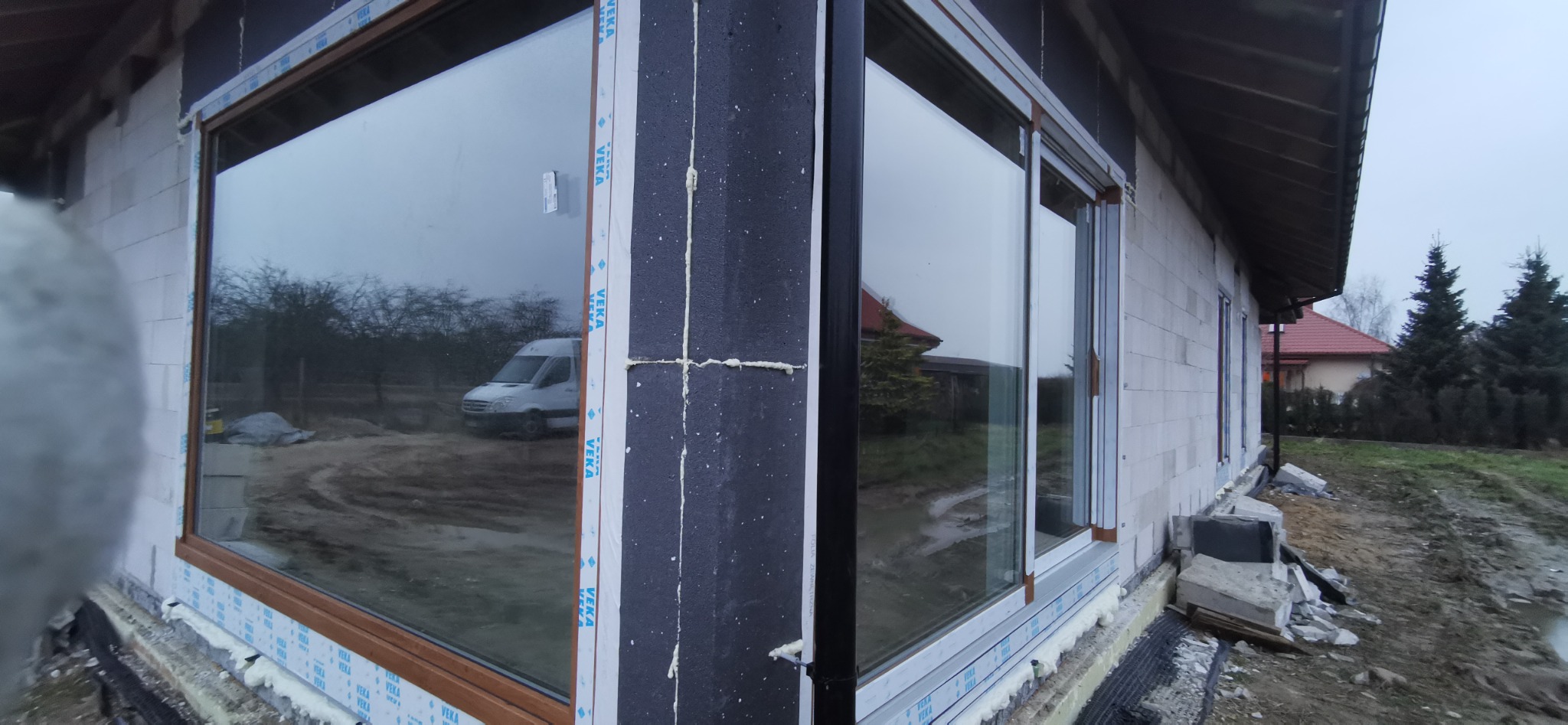 Montaż okien z brązowymi ramami w surowym budynku z białego bloczka, widoczna pianka montażowa i folia zabezpieczająca, w tle zaparkowany biały bus.