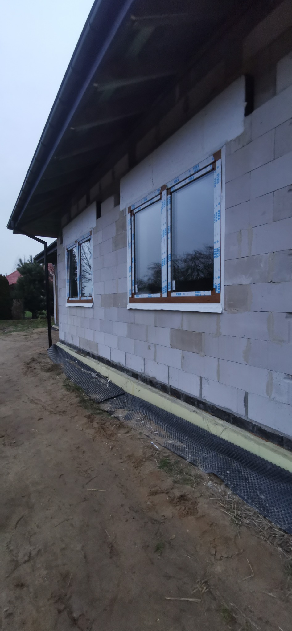 Nowo zamontowane okna w brązowych ramach z folią ochronną na białej elewacji budynku z betonu komórkowego w trakcie budowy, widok z zewnątrz.