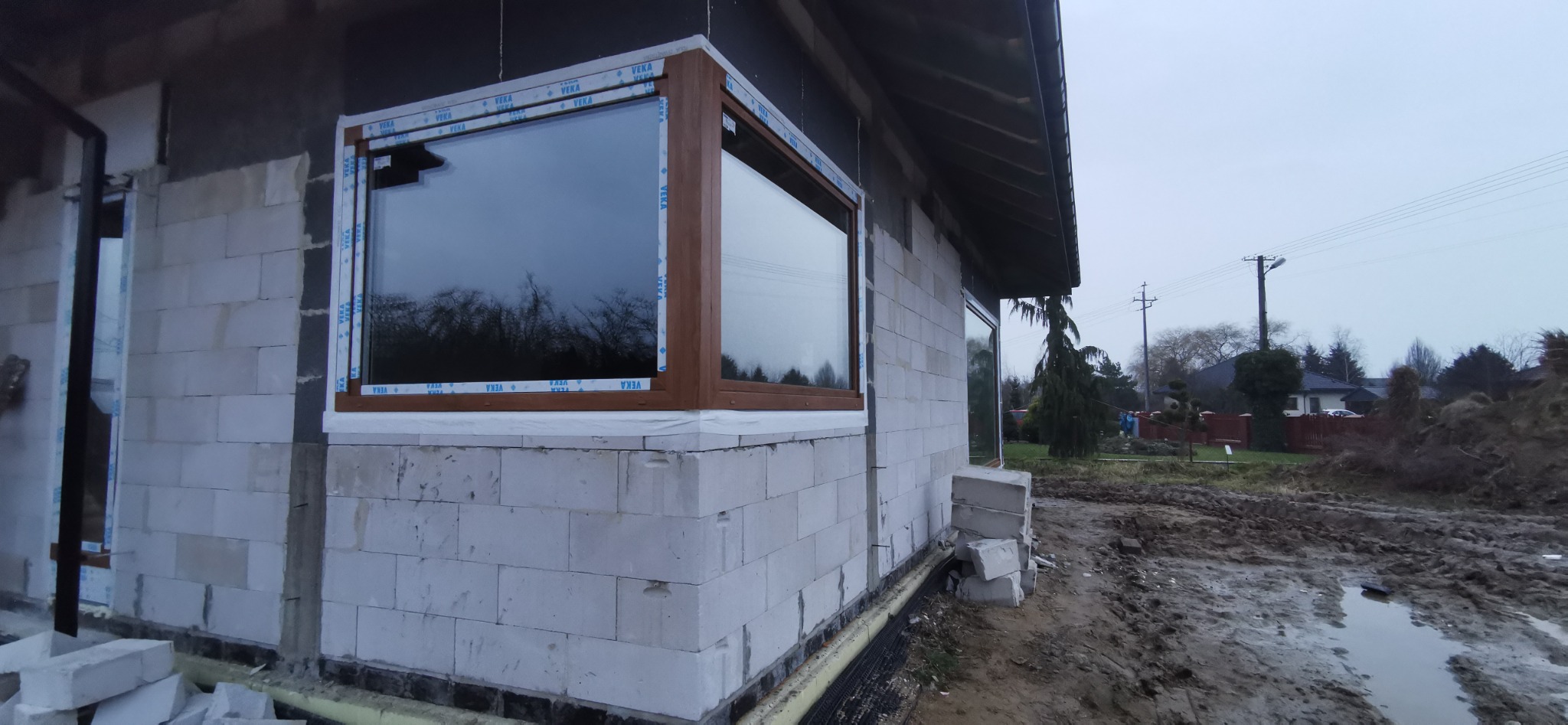 Montaż okna z brązową ramą w surowym stanie budynku z białych bloczków, widoczne folie ochronne na oknach.