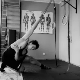 Czarno-białe zdjęcie mężczyzny w sportowym stroju rozciągającego się w sali gimnastycznej z drabinkami, piłką do ćwiczeń i plakatami anatomicznymi na ścianie.