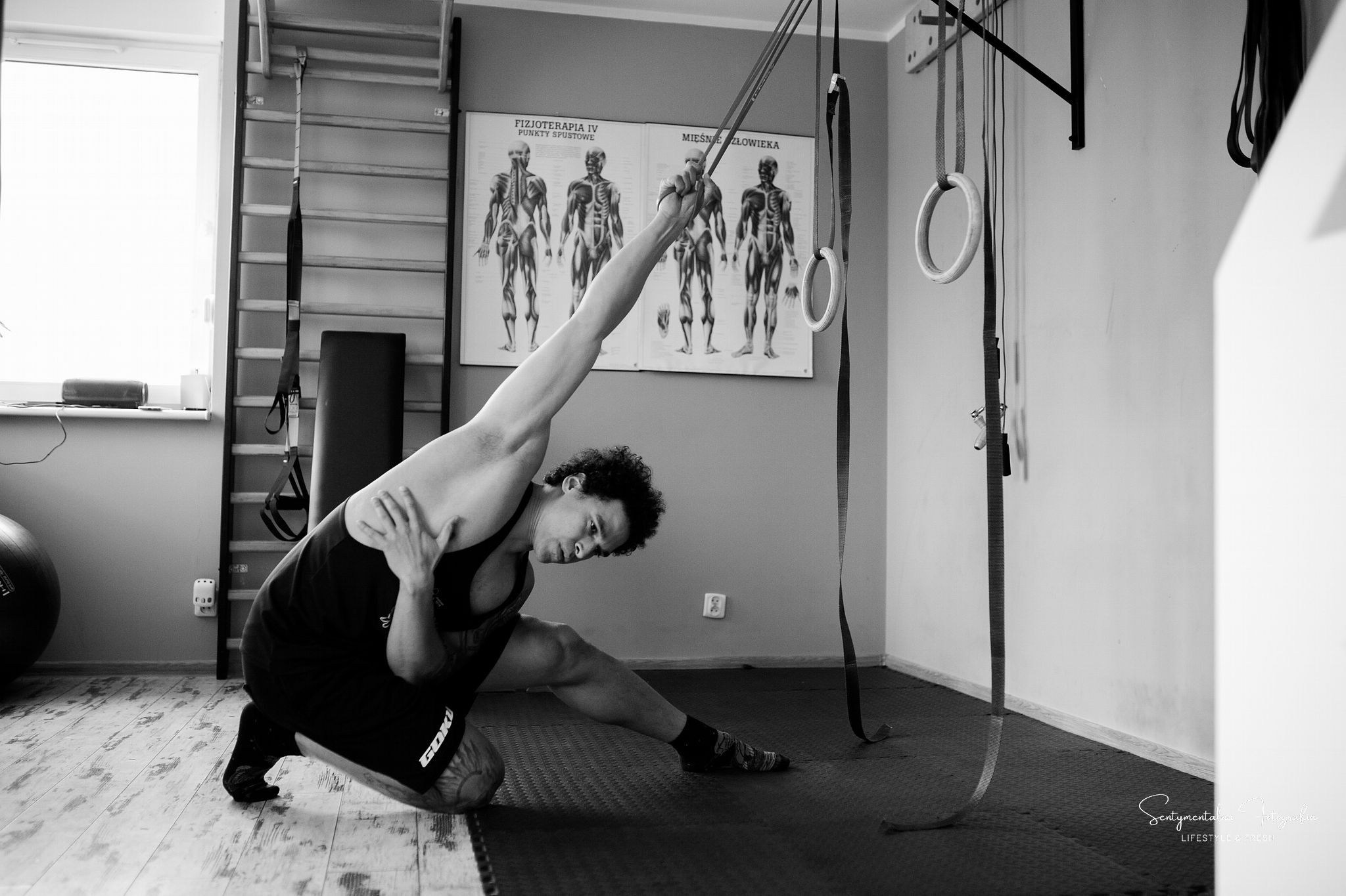 Czarno-białe zdjęcie mężczyzny w sportowym stroju rozciągającego się w sali gimnastycznej z drabinkami, piłką do ćwiczeń i plakatami anatomicznymi na ścianie.