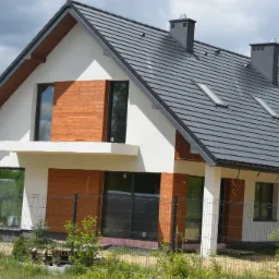 Dwukondygnacyjny dom jednorodzinny z grafitową dachówką i drewnianą elewacją, widoczny zza ogrodzenia z siatki, z zaparkowanym białym vanem w tle.