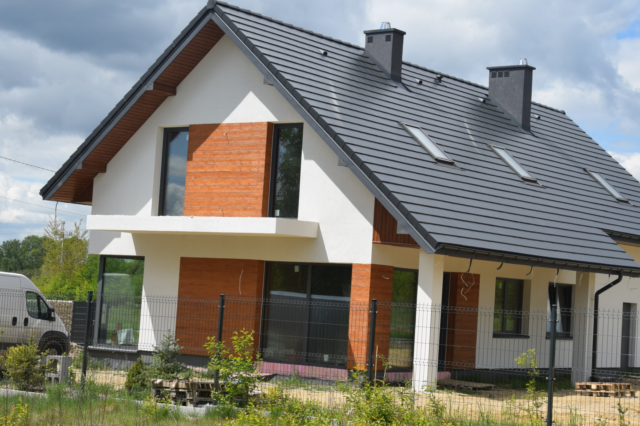 Dwukondygnacyjny dom jednorodzinny z grafitową dachówką i drewnianą elewacją, widoczny zza ogrodzenia z siatki, z zaparkowanym białym vanem w tle.