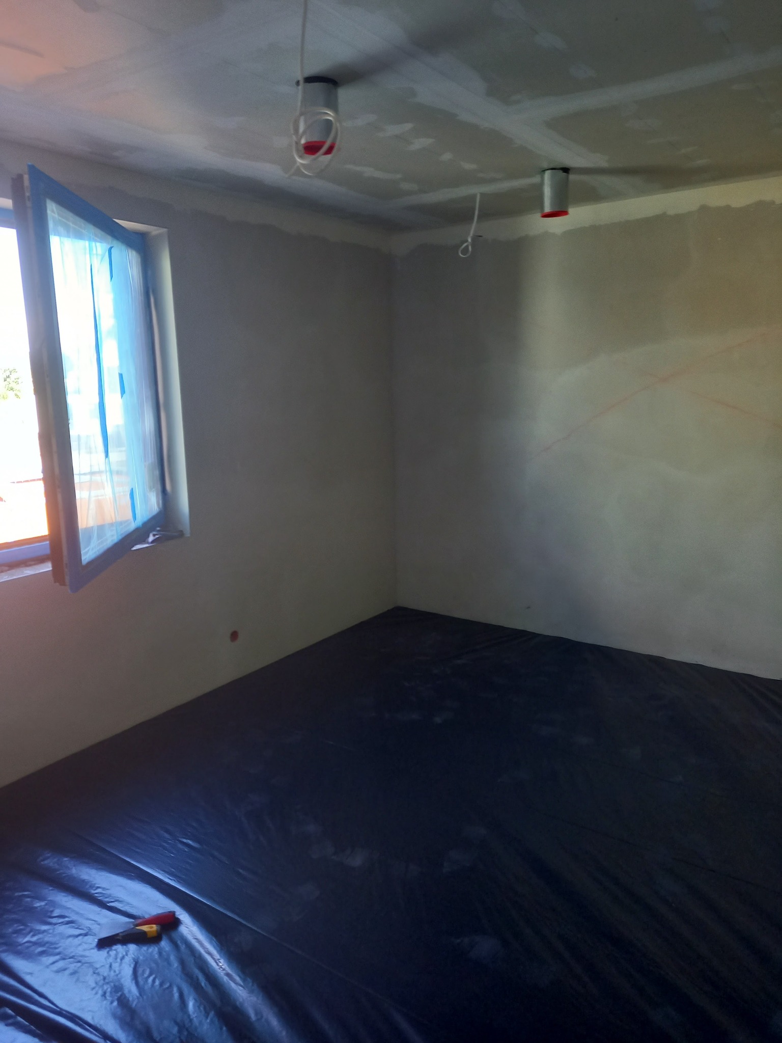 Pokój w trakcie przygotowań do prac wykończeniowych, z zabezpieczoną folią podłogą i oknem oklejonym taśmą malarską, ściany wstępnie przygotowane, widoczne przewody oświetleniowe.