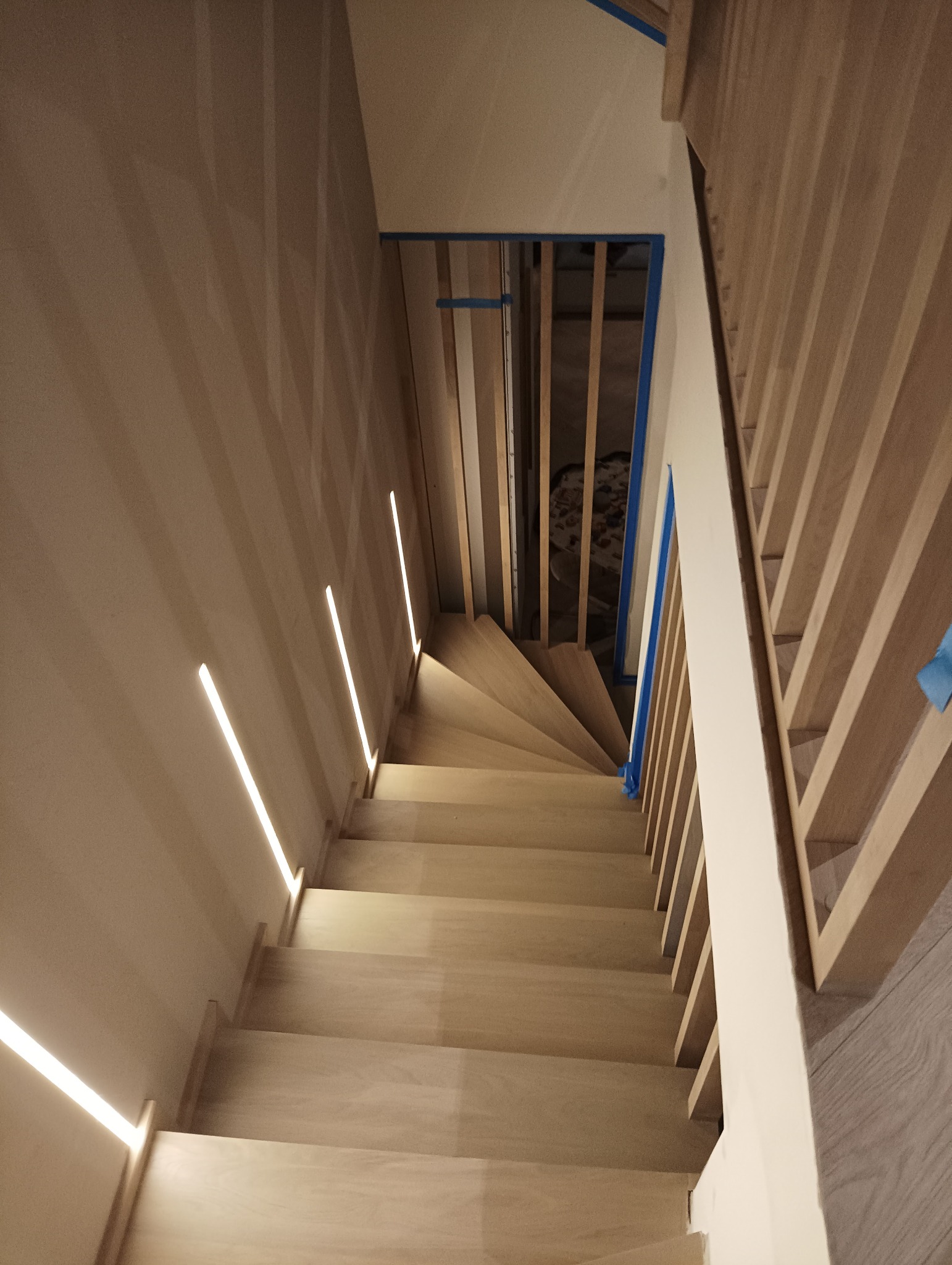 Widok z góry na drewniane schody z podświetleniem LED w ścianie i zabezpieczonymi taśmą malarską elementami balustrady