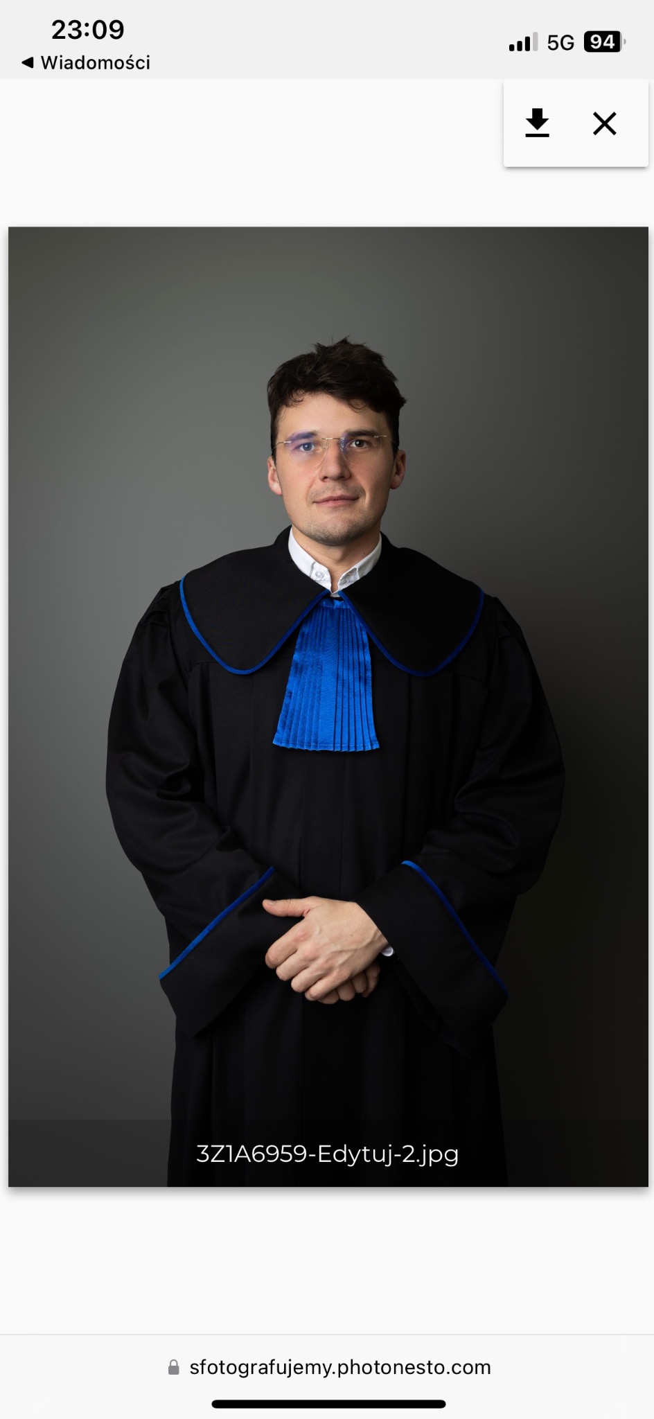 Portret mężczyzny w todze z niebieskim żabotem i okularach, na szarym tle, z rękami splecionymi na wysokości pasa.