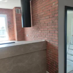 Częściowo wykończona kuchnia z ceglaną ścianą, nowoczesnym okapem i blatem z imitacji kamienia. Widoczny fragment otwartych drzwi i drabina w tle.