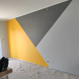 Ściana pomalowana w geometryczne wzory w odcieniach żółtego, szarego i białego, widoczne gniazdka elektryczne i zabezpieczona folią podłoga w trakcie prac wykończeniowych.