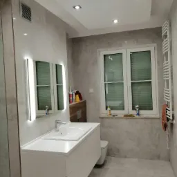 Jasna łazienka po remoncie z umywalką nablatową, podświetlanym lustrem, grzejnikiem drabinkowym i oknem z żaluzjami, wykończona płytkami imitującymi kamień.