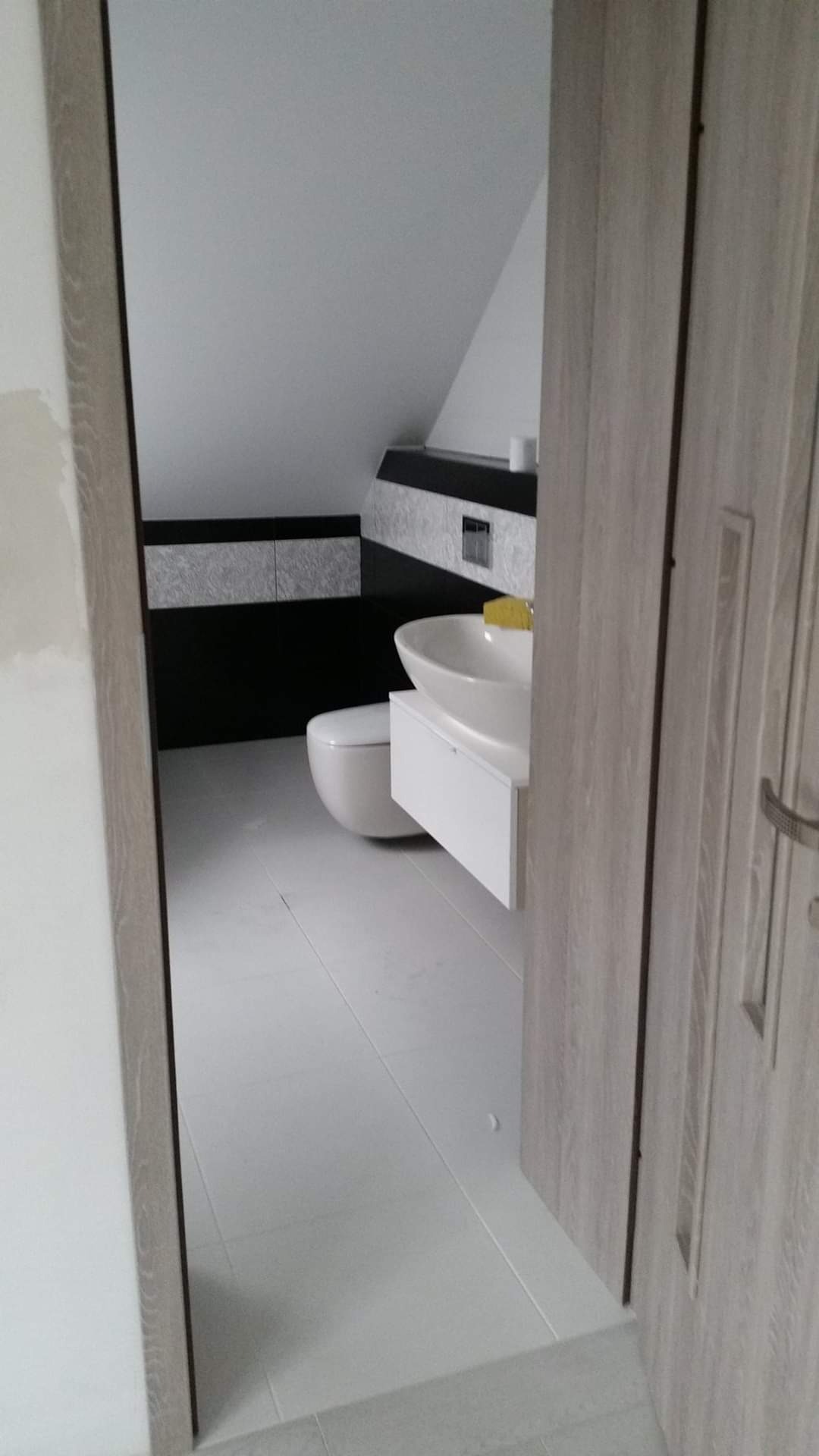 Nowoczesna łazienka na poddaszu z białą ceramiką i czarno-białymi płytkami dekoracyjnymi, widoczna przez częściowo otwarte drzwi z drewnopodobnym wykończeniem.