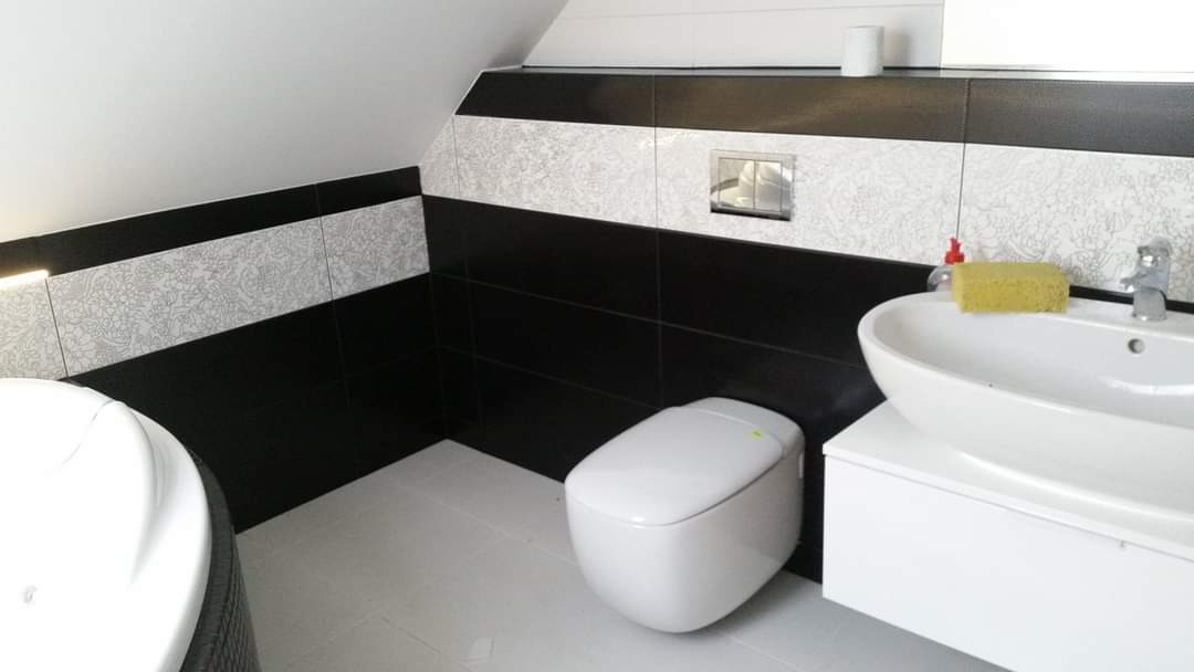 Nowoczesna łazienka z białą umywalką nablatową, podwieszaną toaletą i czarno-białymi płytkami z delikatnym wzorem kwiatowym, oświetlenie LED nad płytkami.