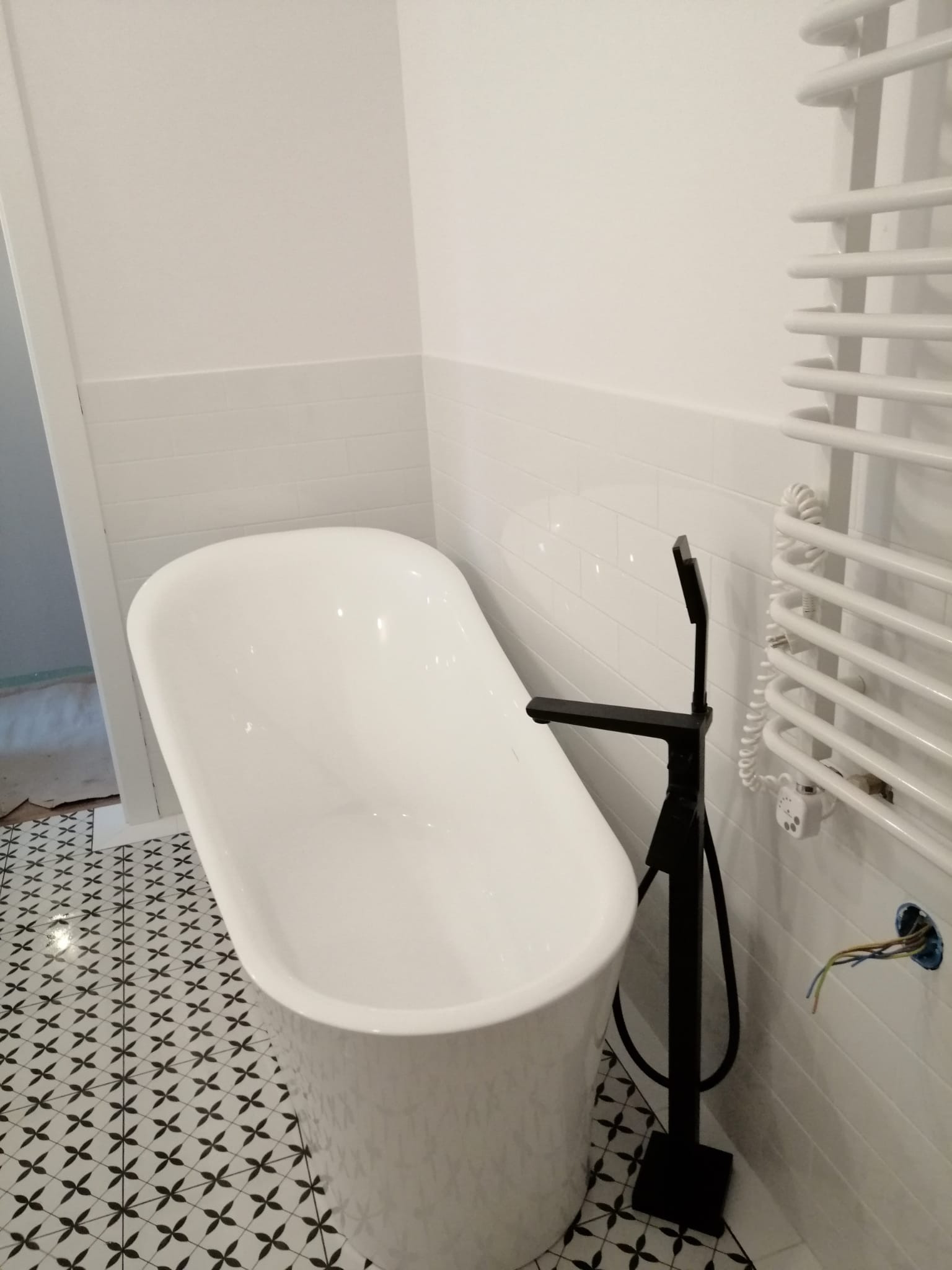 Wolnostojąca wanna w nowocześnie wykończonej łazience z czarno-białą podłogą w geometryczny wzór, czarna bateria podłogowa, biały grzejnik drabinkowy i ściany wyłożone białymi, prostokątnymi płytkami.