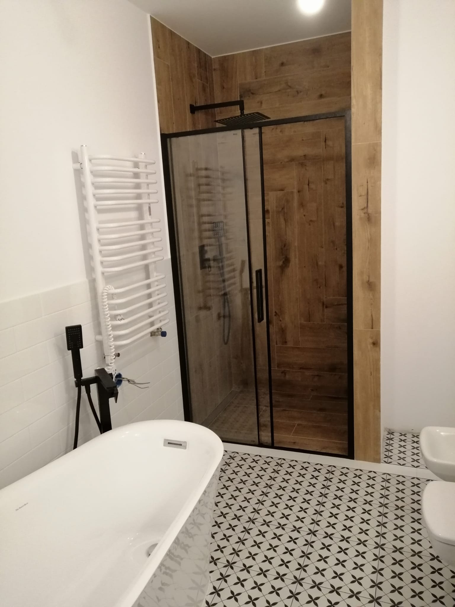 Nowoczesna łazienka z kabiną prysznicową w czarnej ramie, drewnopodobnymi płytkami na ścianie prysznica, białą wanną wolnostojącą i grzejnikiem drabinkowym na białej ścianie, oraz wzorzystą podłogą.