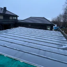 Montaż blachy na rąbek stojący na nowym dachu, widoczne mocowania i zielona mata pod spodem, w tle budynek z ciemną elewacją i dachem.