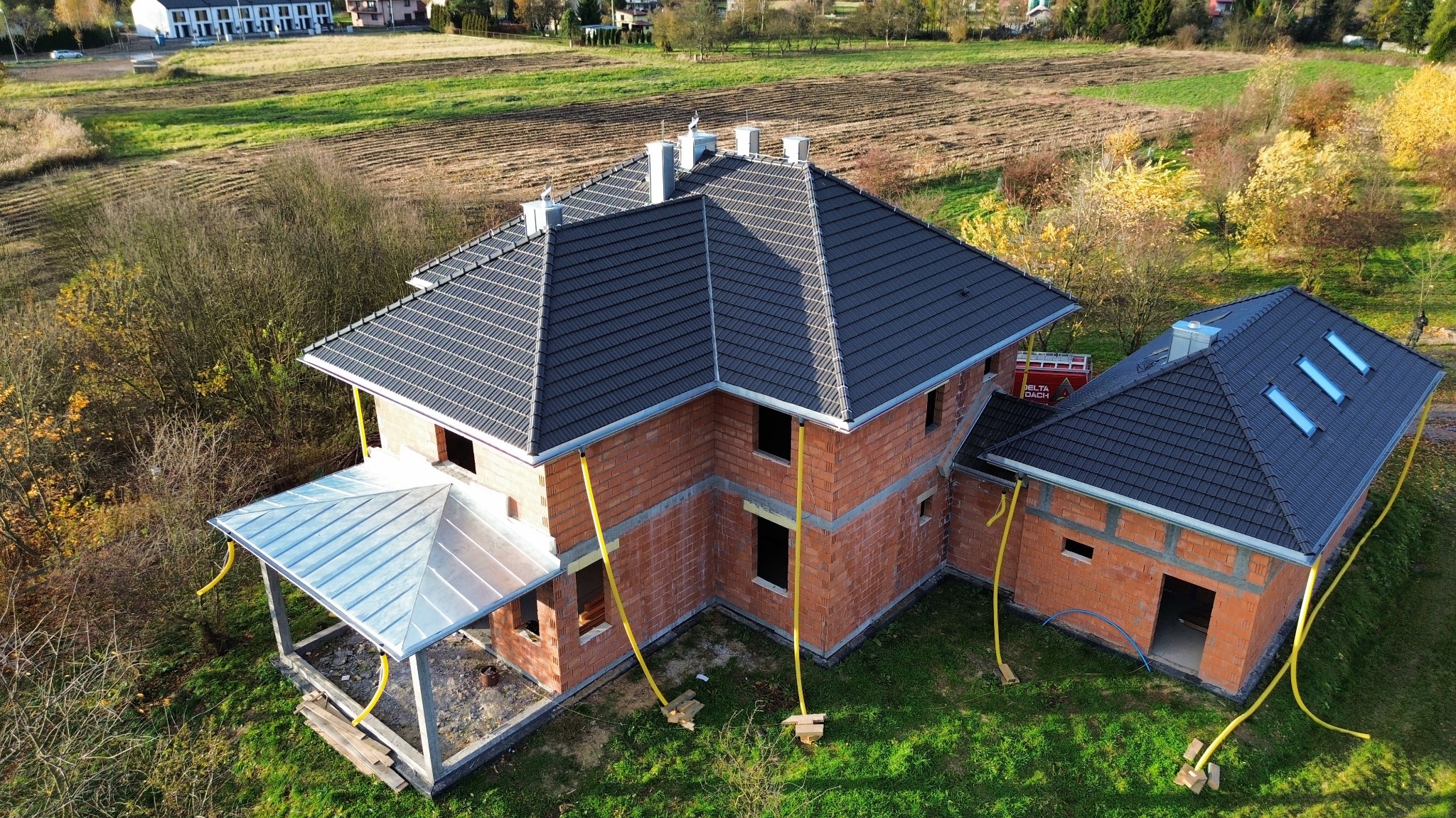 Dom w trakcie budowy z ciemną dachówką, widok z góry. Na dachu okna dachowe. Czerwona cegła, żółte rury spustowe. W tle pole i zabudowania.