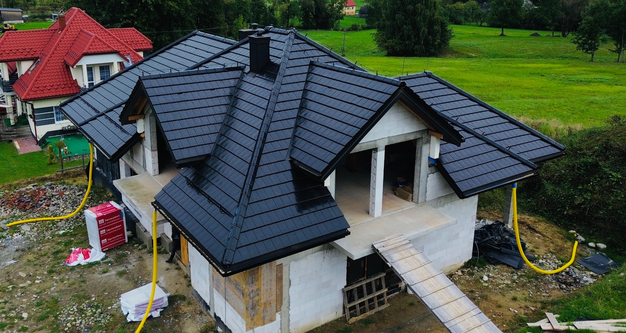 Nowoczesny dom w budowie z czarnym, błyszczącym dachem z dachówki ceramicznej. Widok z góry na częściowo ukończony budynek i zielone otoczenie.
