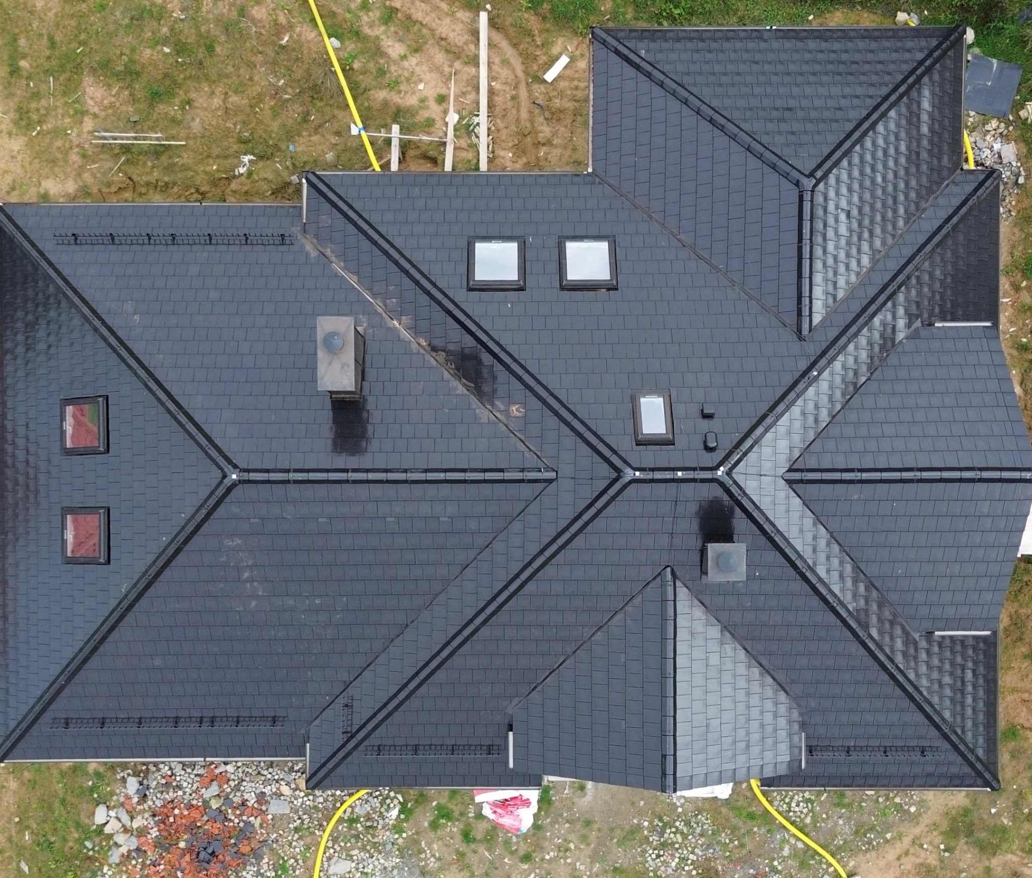 Widok z góry na ciemny, symetryczny dach z dachówki, z oknami dachowymi i kominem. Wokół dachu widoczny teren z gruzem i wężem ogrodowym.