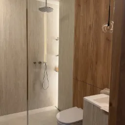 Nowoczesna łazienka z kabiną prysznicową typu walk-in, toaletą i drewnianą zabudową. Minimalistyczny design, jasna kolorystyka, eleganckie oświetlenie.